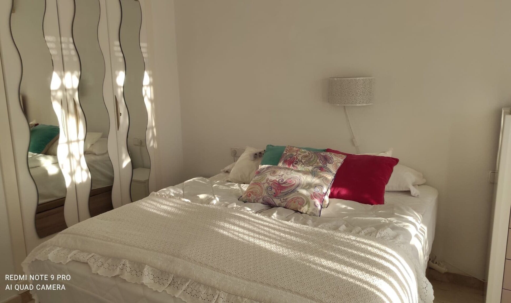 Una cama bien tendida con edredón blanco y almohadones decorativos, una lámpara de noche y un espejo con marco ondulado.