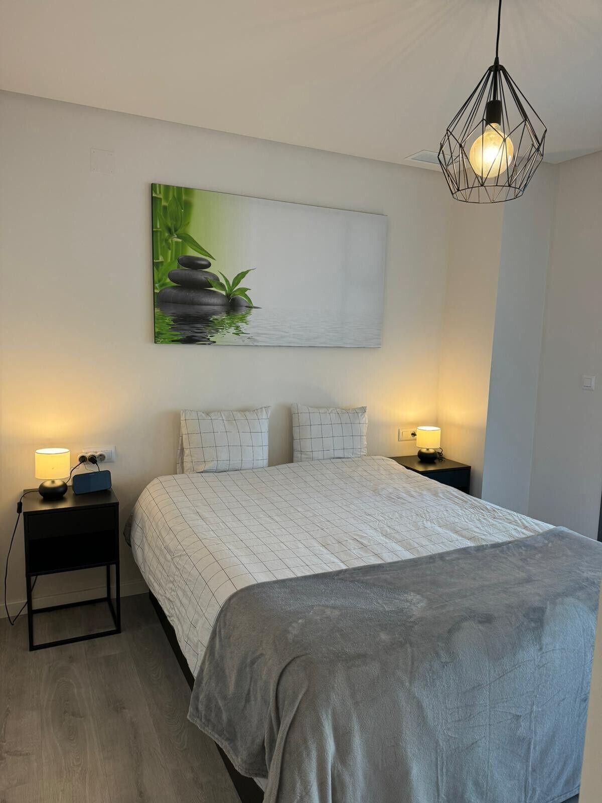 Ein Schlafzimmer mit einem Bett, zwei Nachttischen, einer Wandleuchte und einem Bild von Steinen und Pflanzen.