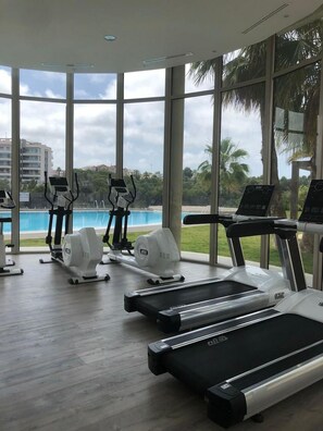 Un gimnasio con cintas de correr, elípticas y piscina, que cuenta con amplias ventanas y vistas a palmeras y edificios.