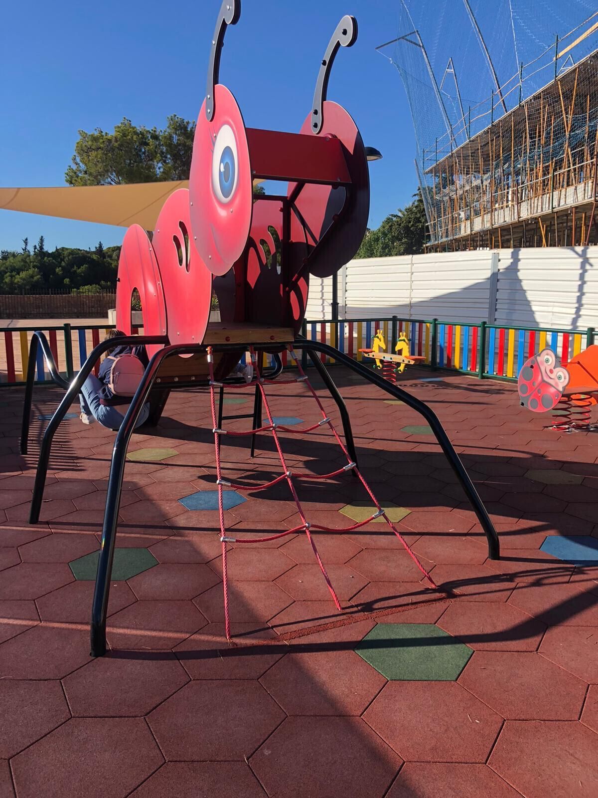 Une aire de jeux avec une grande structure d’escalade en forme de fourmi rouge, entourée d’une clôture colorée et d’une zone délimitée par un filet à l’arrière-plan.