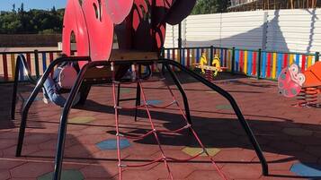 Une aire de jeux avec une grande structure d’escalade en forme de fourmi rouge, entourée d’une clôture colorée et d’une zone délimitée par un filet à l’arrière-plan.