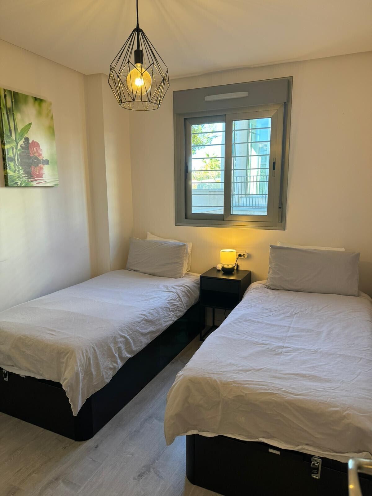 A bedroom with two beds, a nightstand with a lamp, a window with blinds, and a hanging light fixture.