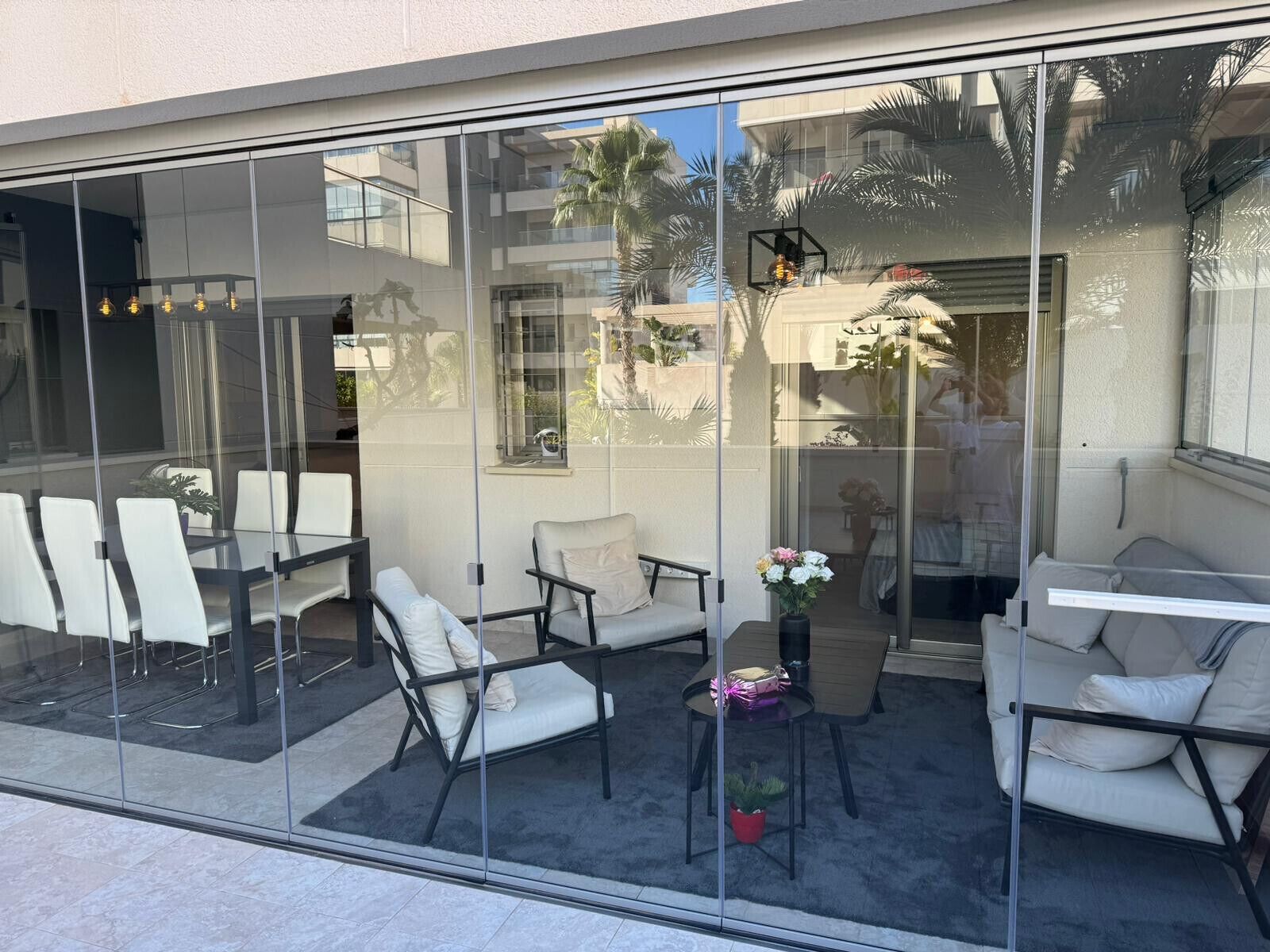 A modern outdoor seating area with glass walls, white chairs, and a small table with flowers.