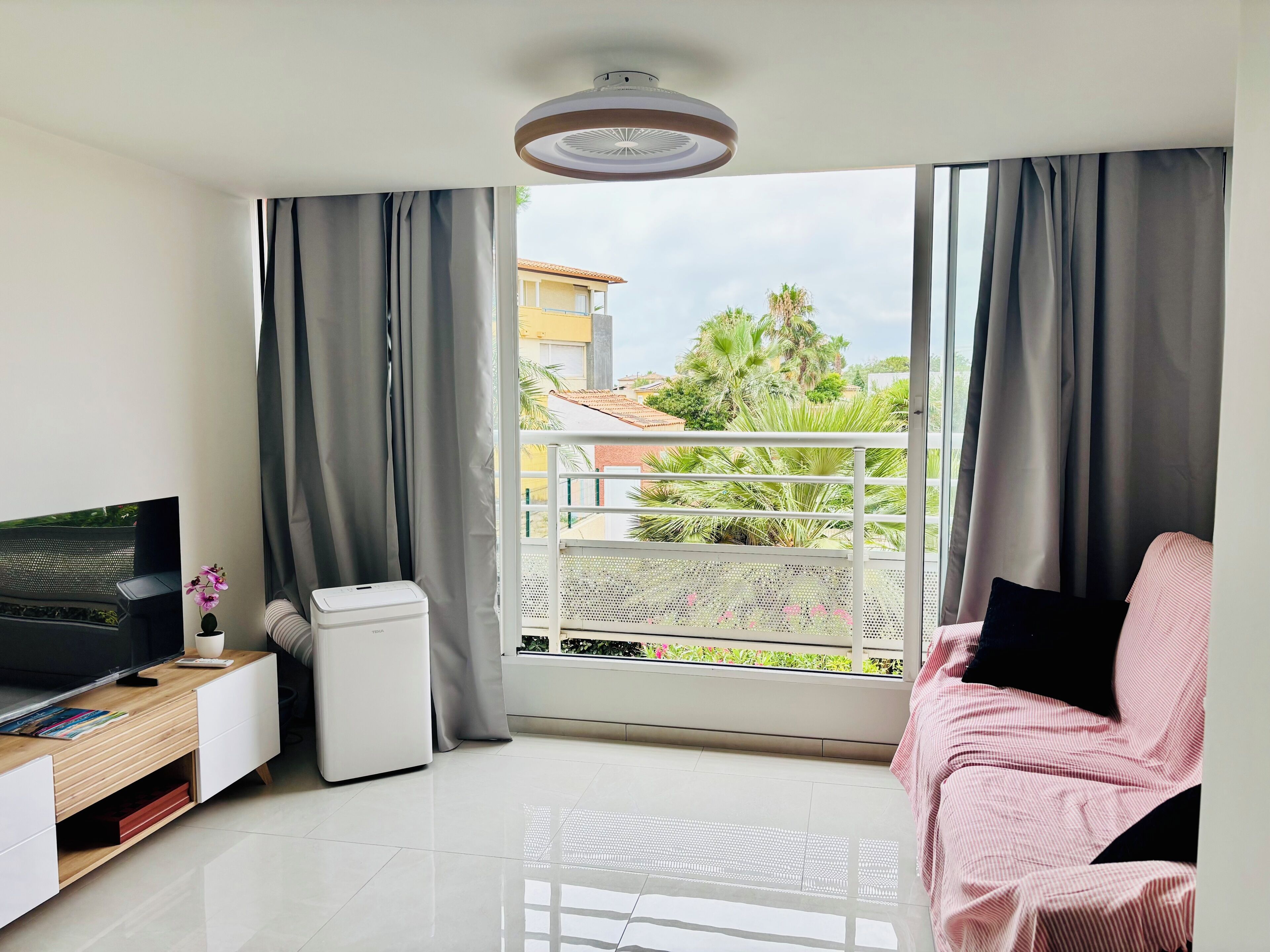 Ein modernes Wohnzimmer mit Flachbildfernseher, einem kleinen weißen Gerät, einem Holznachttisch, einem rosa Sessel und einem Balkon mit Blick auf Gebäude und Grünflächen.