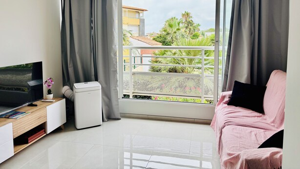 Ein modernes Wohnzimmer mit Flachbildfernseher, einem kleinen weißen Gerät, einem Holznachttisch, einem rosa Sessel und einem Balkon mit Blick auf Gebäude und Grünflächen.