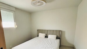 Ein kleines Zimmer mit einem Einzelbett, einer neutralen Farbgestaltung, einem Fenster mit Jalousien und einem Deckenventilator.