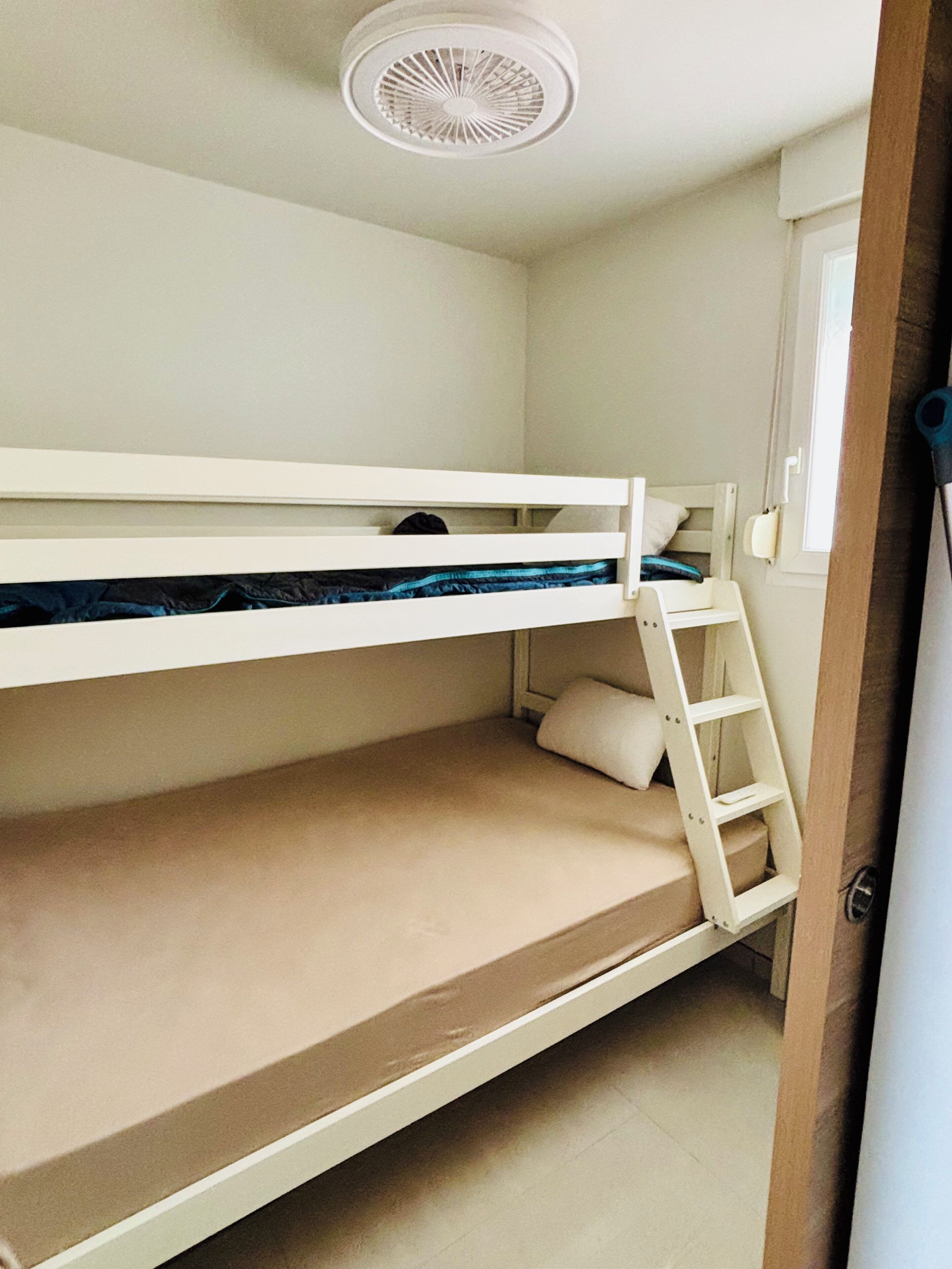 Una cama litera con escalera, un ventilador de techo y una ventana con cortina.