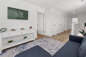Ein modernes Wohnzimmer mit einem grauen Sofa, einem an der Wand befestigten Fernseher, der 'ROYAL STAYS' anzeigt, und einem Flur, der zu einer Tür führt.