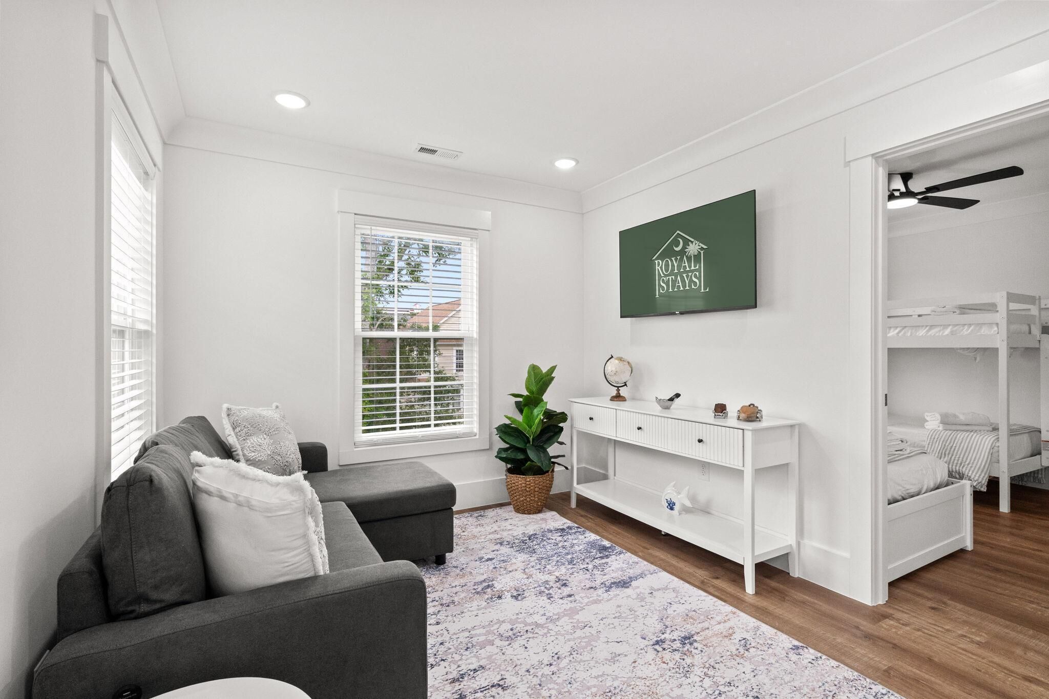 A modern living room with a grey sofa, a white console table, a potted plant, a bunk bed, and a large window with blinds.