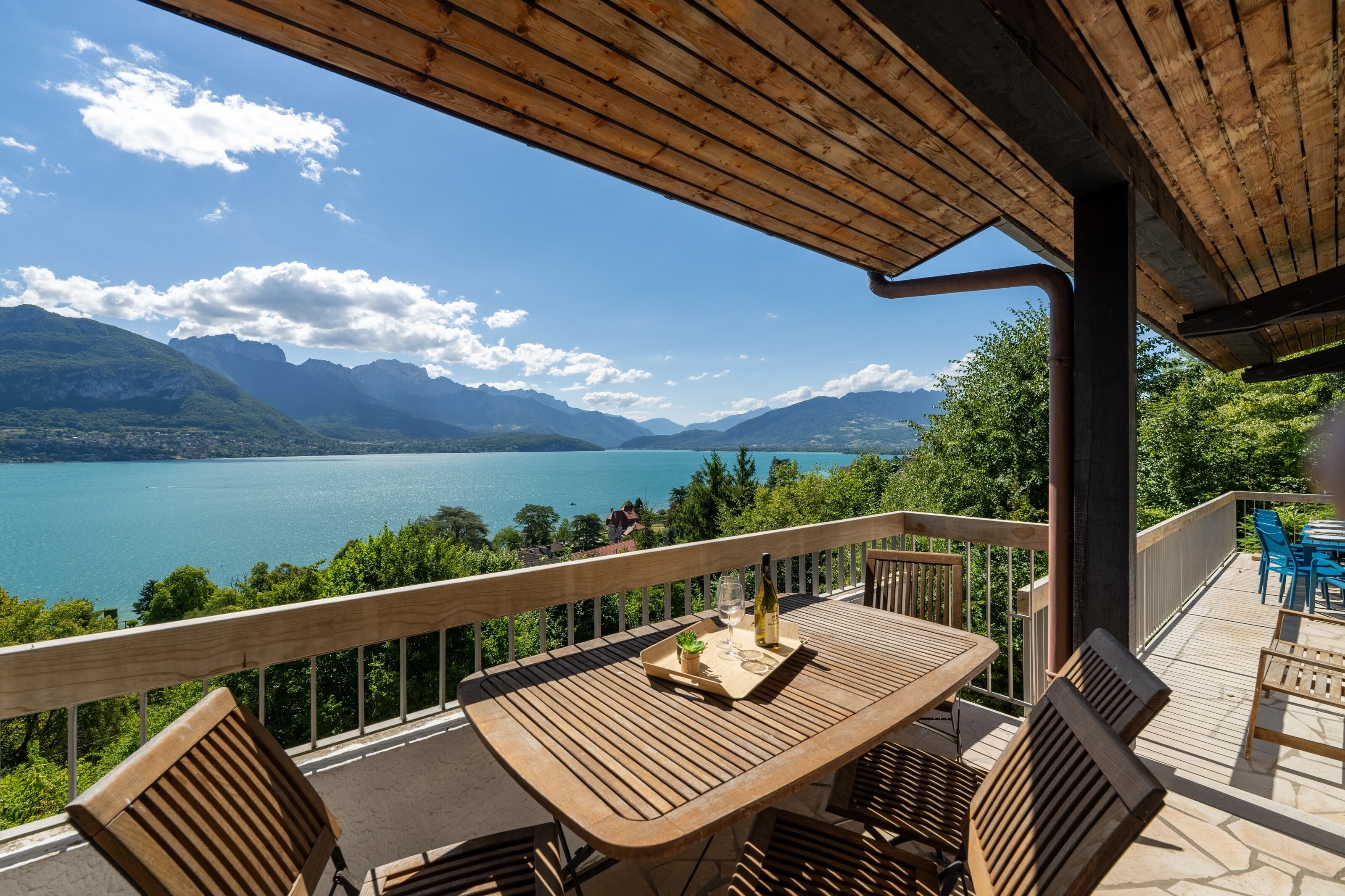 A wooden deck with a table and chairs, overlooking a lake and mountains.