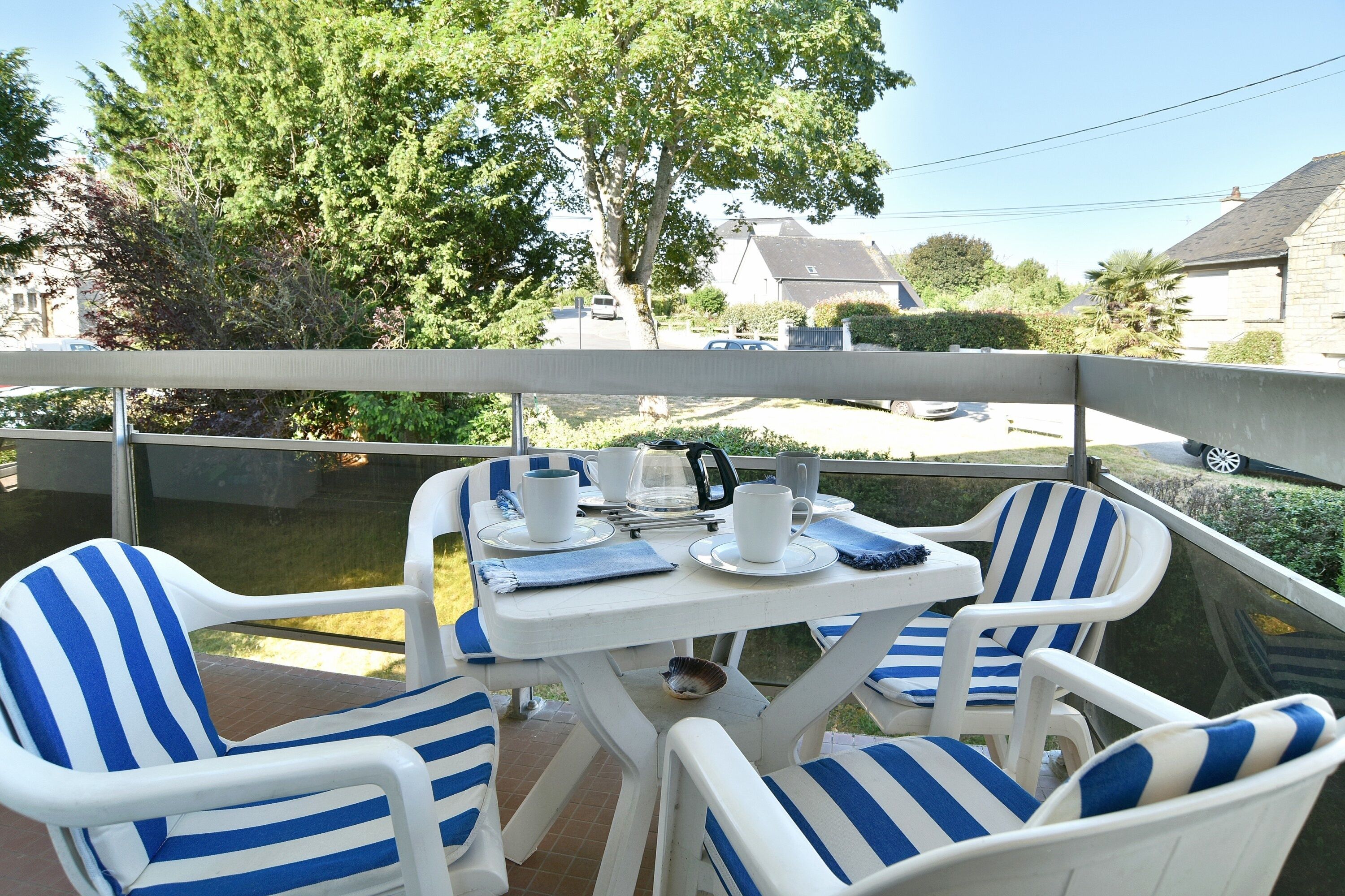 Un balcon avec une table dressée pour deux personnes, deux chaises à rayures et une vue sur la rue et les arbres.