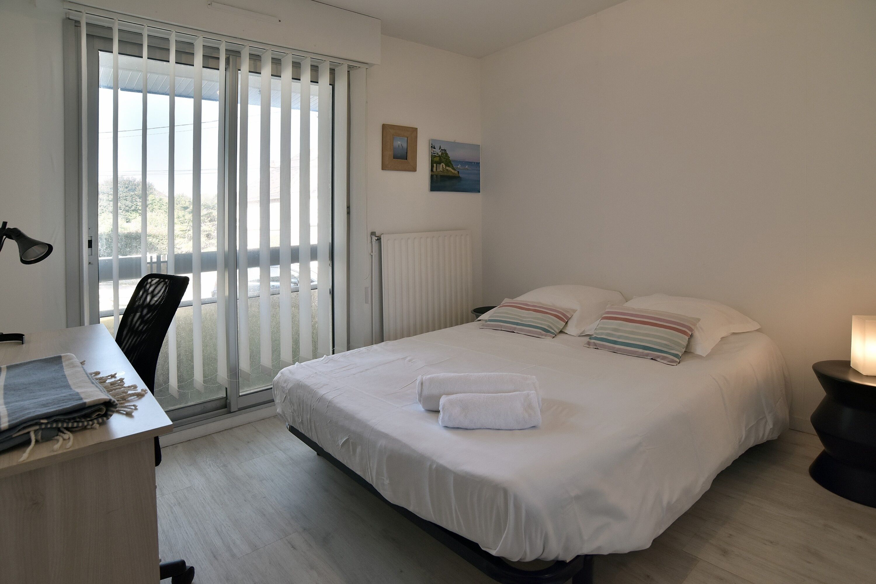 A bedroom with a bed, a desk, a chair, a radiator, and a window with blinds.