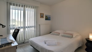 A bedroom with a bed, a desk, a chair, a radiator, and a window with blinds. - Le Balcon d'Émeraude - À moins d’1 km de la plage (Dinard)
