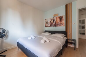 A neatly made bed with towels, a bedside table, a wall-mounted fan, and a door leading to another room. - Perle de Strasbourg - Bel appt pour 4 avec balcon (Strasbourg)