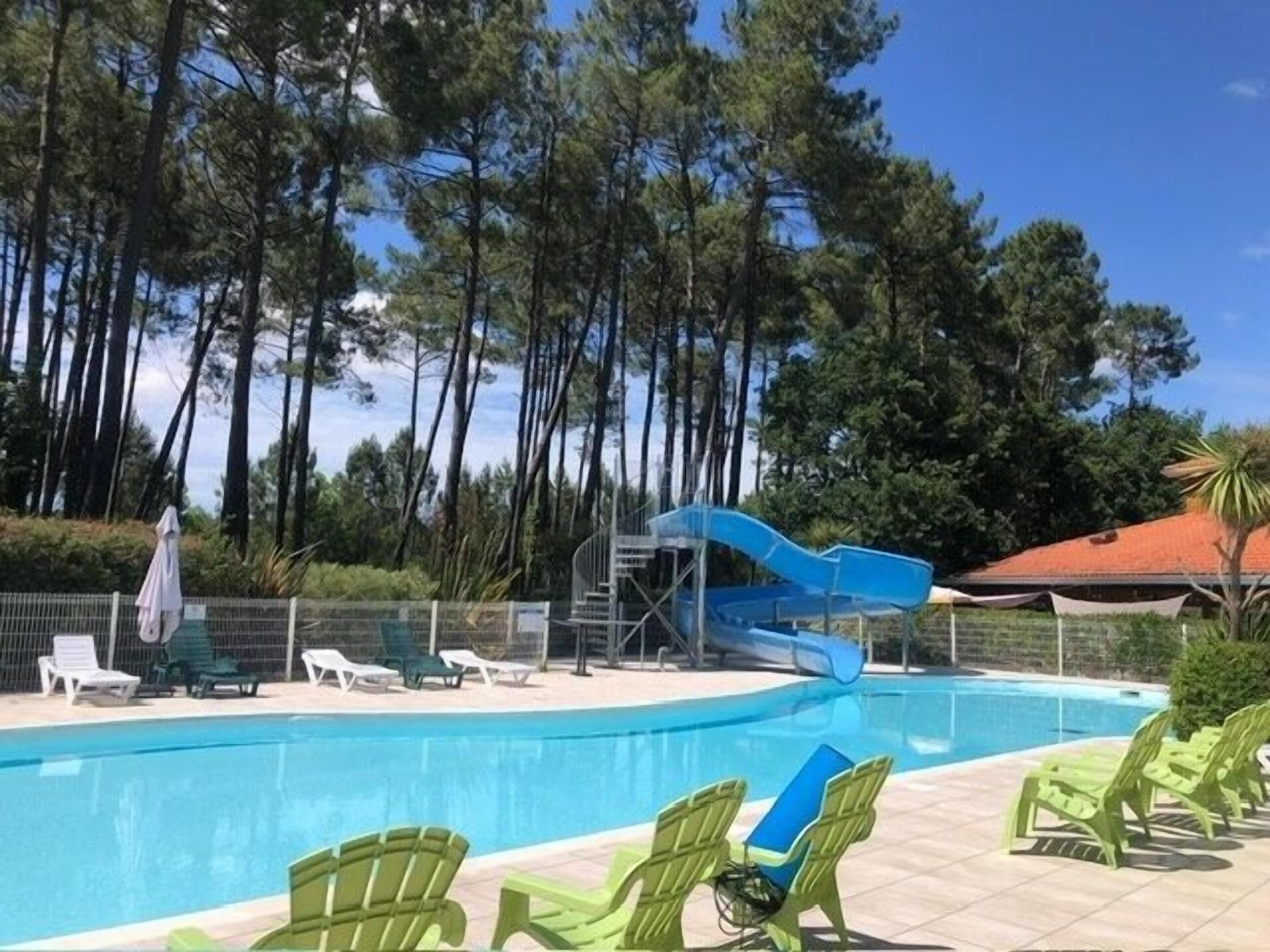 Une piscine avec un toboggan bleu, des chaises longues vertes et un espace clos avec des arbres.