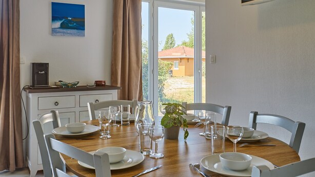 Zona pranzo con tavolo in legno apparecchiato per quattro persone, sedie bianche, una pianta in vaso e vista attraverso una porta vetrata.