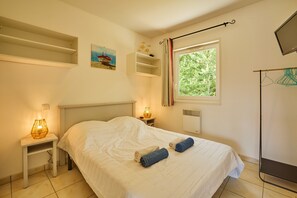 Ein Schlafzimmer mit Bett, Nachttisch, Lampe, Fenster mit Blick auf Bäume und einem Regal mit Dekoration.