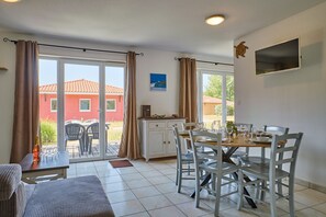 A dining area with a table set for four, a sofa, and a kitchen area with a refrigerator and a coffee machine. - La Parenthèse Bleue - À 5 min à pied de la plage (Parentis-en-Born)