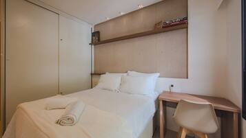 A neatly made bed with a rolled towel, a wooden desk, and a chair in a modern room with a headboard and a shelf with books.