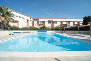 Ein Swimmingpool mit klarer, blauer Wasseroberfläche, umgeben von einem weiß gefliesten Deck. Im Hintergrund ein mehrgeschossiges Gebäude mit Balkonen und einer flachen Dachterrasse.