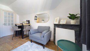 Uma sala de estar com um sofá cinzento, dois bancos altos, um tapete branco, uma mesa lateral verde, uma lareira e um espelho grande.