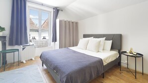 Quarto com cama, mesa de cabeceira, radiador, janela com cortinas e vista para prédios.