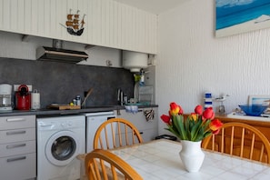 A compact kitchen with a washing machine, a coffee maker, and a dining area with a vase of tulips. - Le Petit Nid de Saint Pierre - À 100 m de la plage (Fleury)
