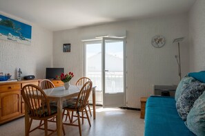 A room with a dining table, chairs, a television, a sofa, and a window with a view. - Le Petit Nid de Saint Pierre - À 100 m de la plage (Fleury)