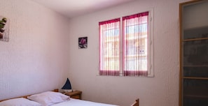 A bedroom with a bed, bedside table, lamp, window with curtains, and a small picture on the wall. - Le Petit Nid de Saint Pierre - À 100 m de la plage (Fleury)