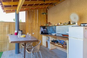 Uma cozinha ao ar livre com pérgola de madeira, mesa, cadeiras e geladeira.