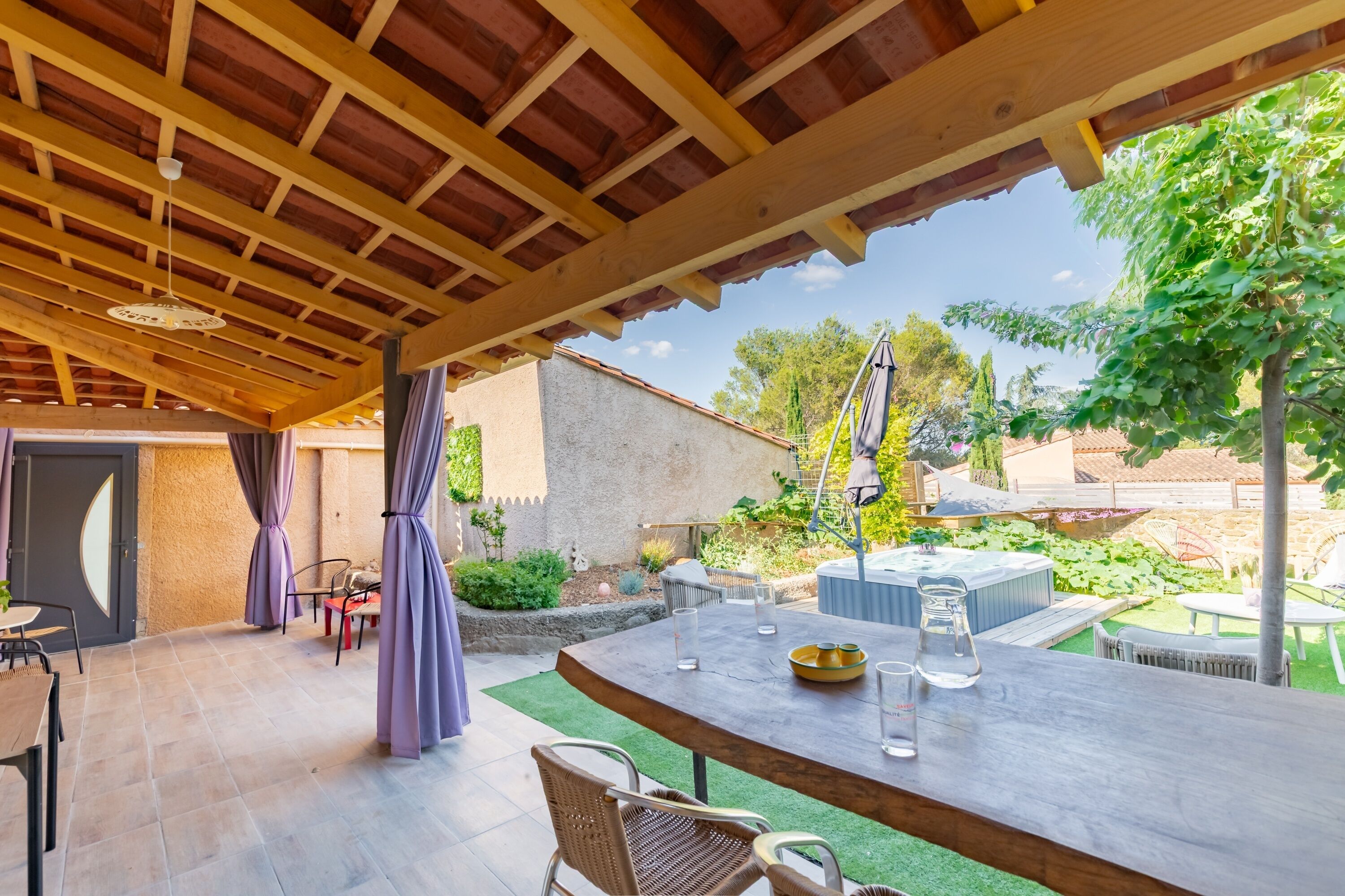 Une terrasse extérieure dotée d’une pergola en bois, d’une table avec des chaises et d’un jacuzzi en arrière-plan.