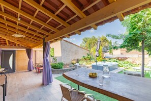 Um pátio externo com pérgola de madeira, mesa com cadeiras e uma banheira de hidromassagem ao fundo.