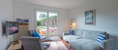 A living room with a sofa, armchair, TV, and a view of a garden through a glass door.