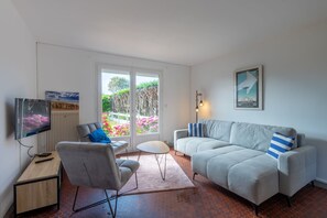 Een woonkamer met een bank, fauteuil, televisie en uitzicht op een tuin door een glazen deur.