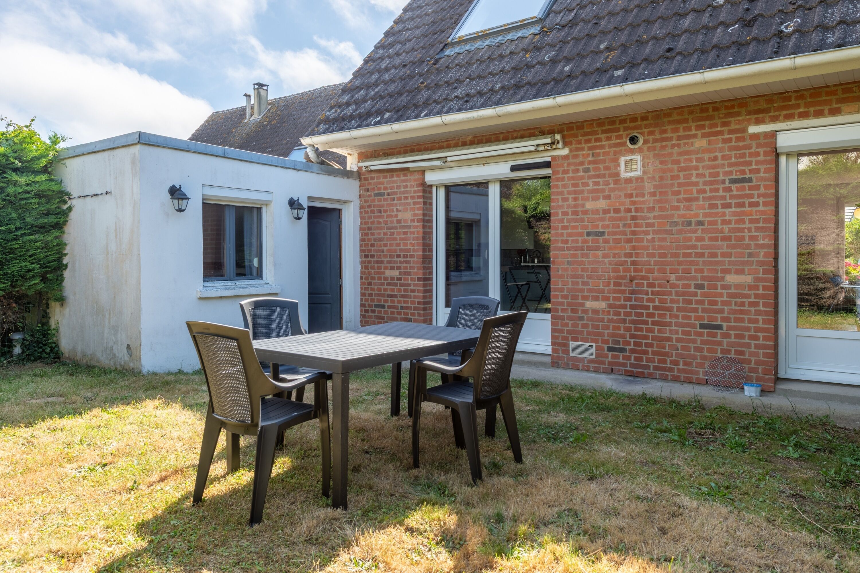 A patio with a table and chairs, a brick house with a sliding glass door, and a small white building.