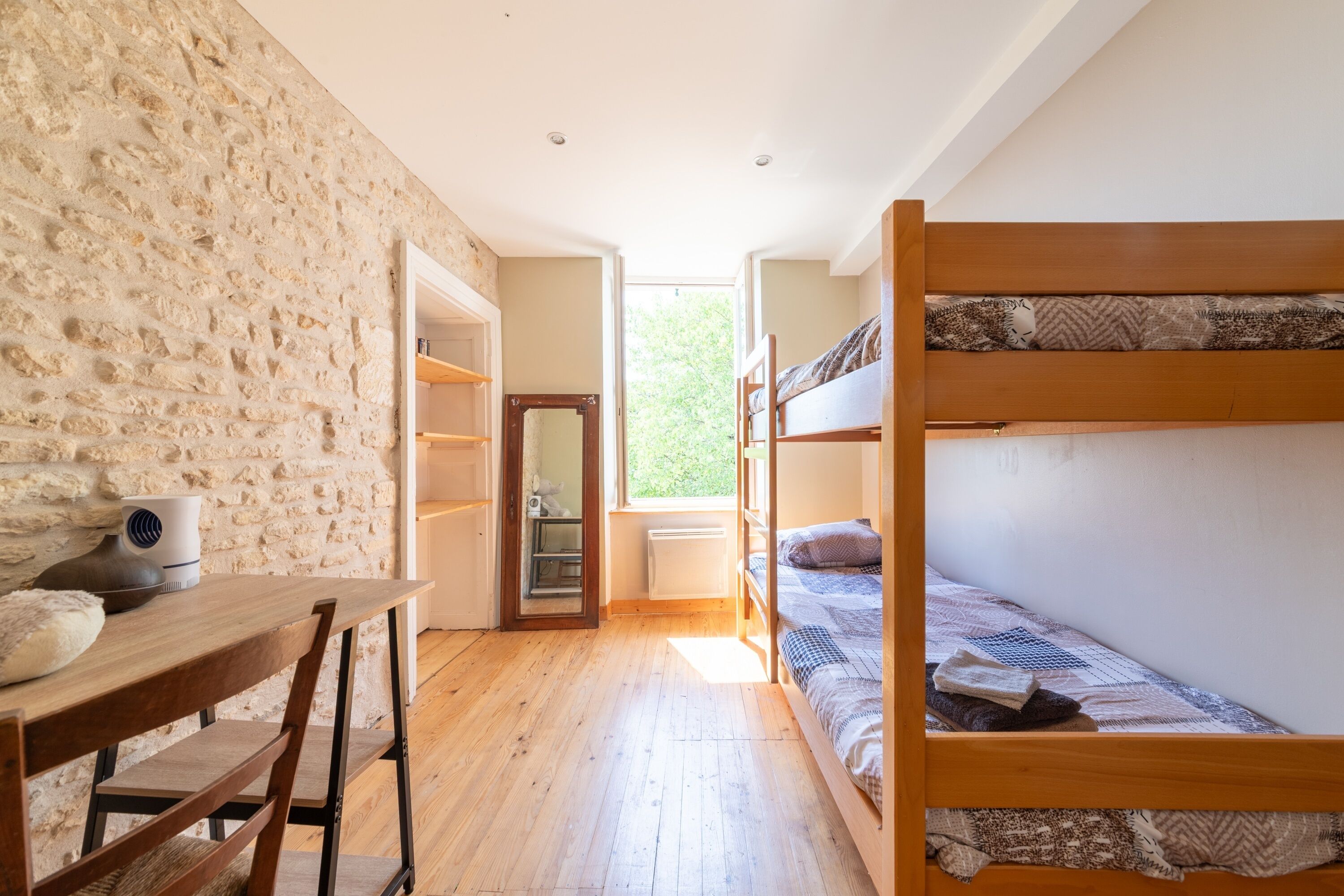 Une pièce avec des lits superposés, une table en bois, un mur en pierre et une vue sur la verdure à l’extérieur.