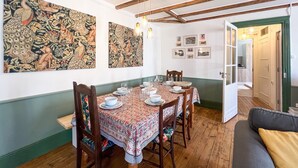 Zona pranzo con tavolo in legno apparecchiato per quattro, circondato da sedie, con un grande arazzo a parete e un'apertura che dà su un'altra stanza.