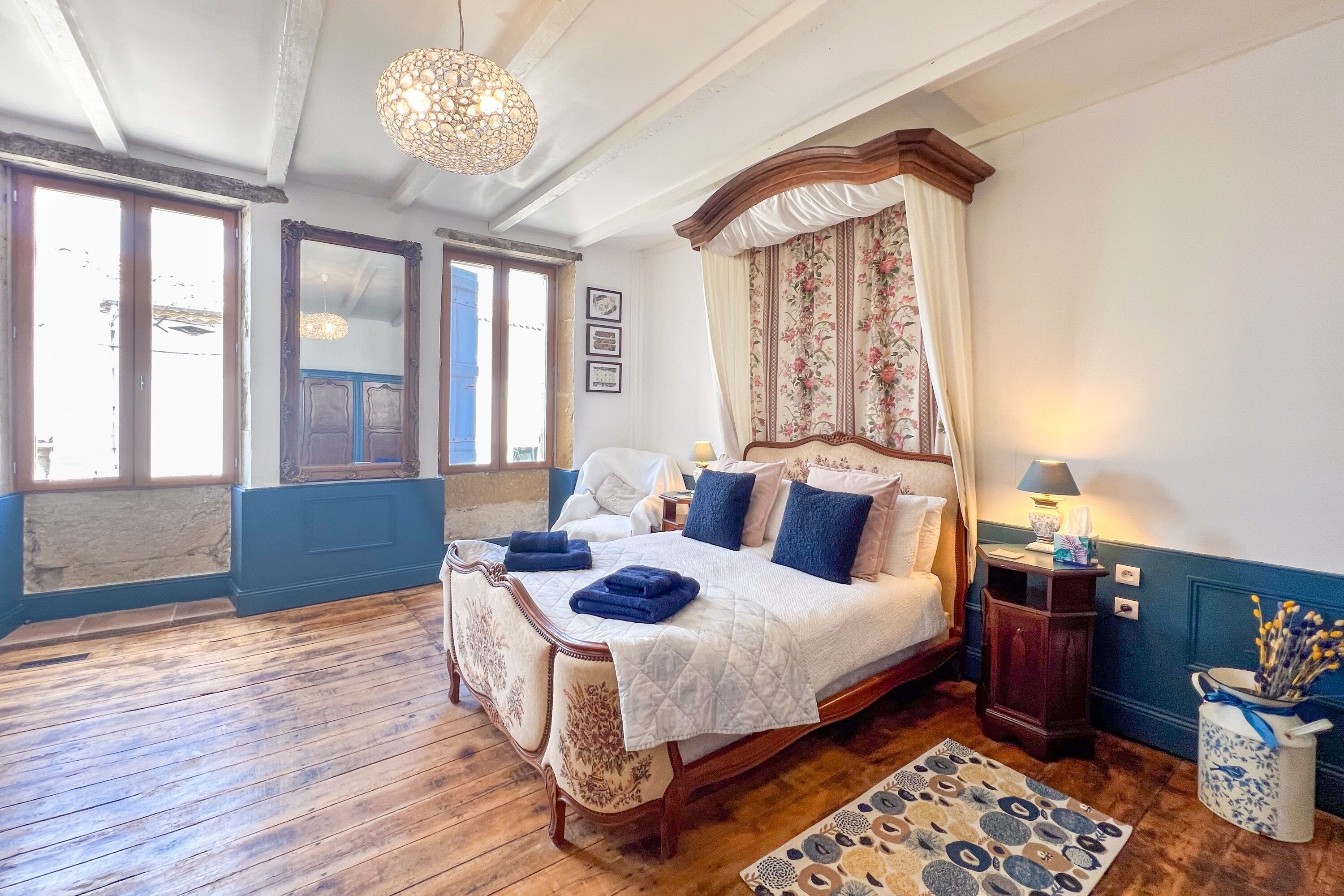 Une chambre à coucher avec un grand lit, un parquet en bois, un lit à baldaquin, un lustre et un vase rempli de fleurs.