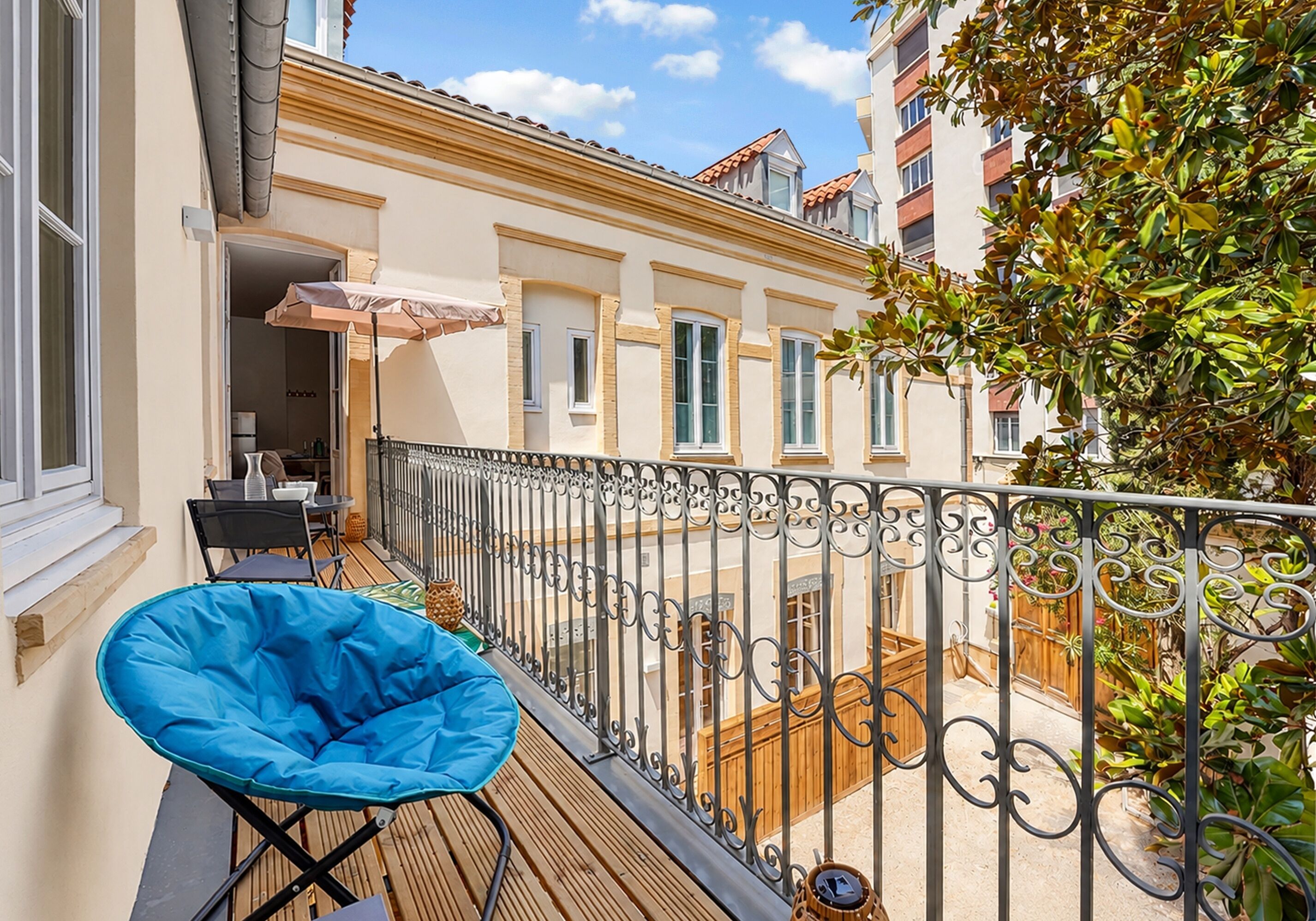 Un balcón con una silla azul, una mesa de madera y vistas a edificios cercanos.