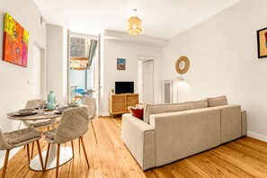 A living room with a sofa, a dining table with chairs, a television, and a hanging light fixture. - La Fabrik Matabiau - Charmant appt avec terrasse et parking (Toulouse)