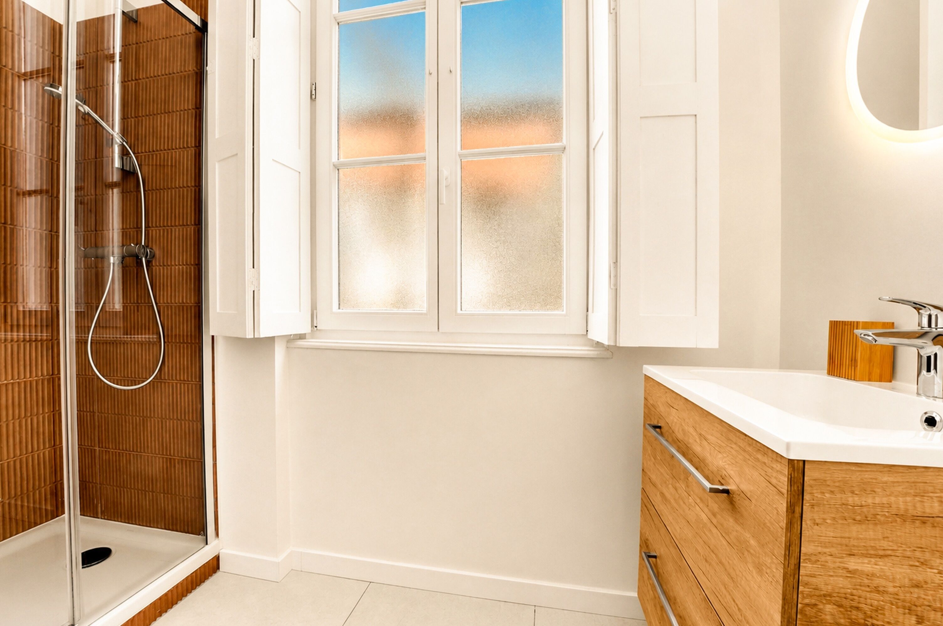 Un baño moderno con cabina de ducha de vidrio, tocador blanco con mueble de madera y ventana esmerilada.