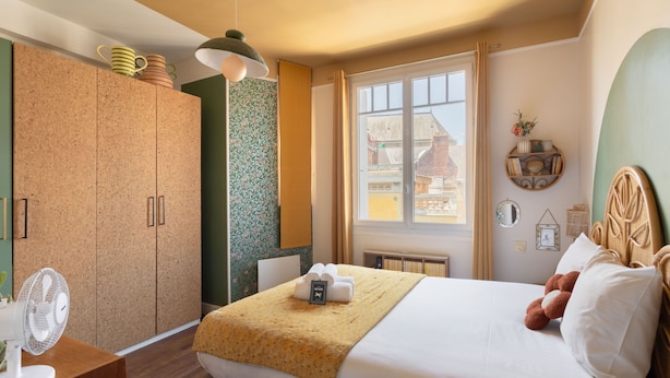Hotellihuone, jossa on sänky, suuri ikkuna, vaatekaappi ja tuuletin.