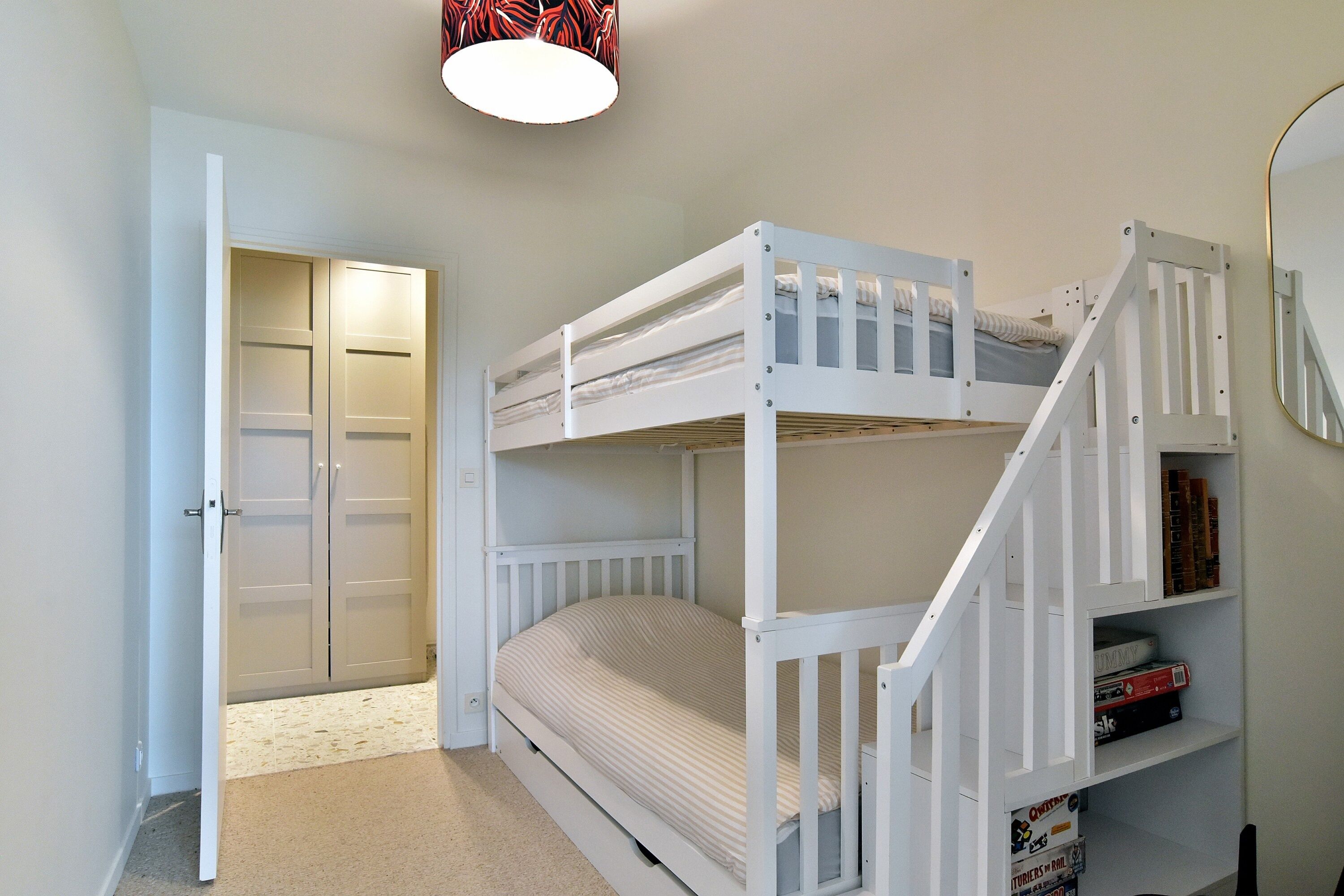 Une chambre avec un lit superposé, une porte, une bibliothèque et un luminaire au plafond.