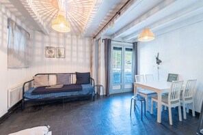 Ein Zimmer mit einem Bett, einem Esstisch mit Stühlen, einem kleinen Hocker und einem Fenster mit Vorhängen.