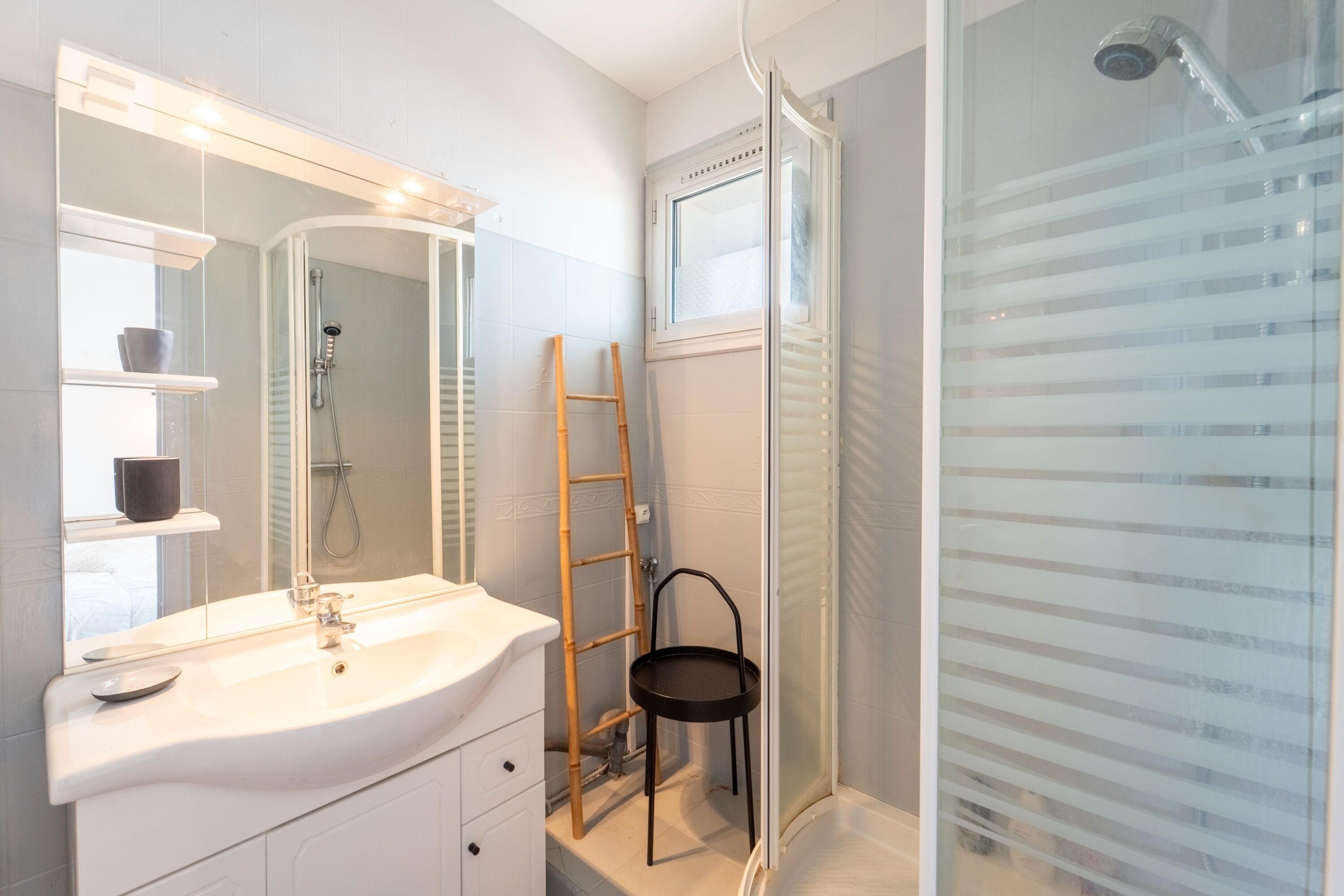 Ein Badezimmer mit einem weißen Waschbecken, einem Spiegel und einer Dusche. Eine Leiter lehnt an der Wand, und ein kleiner Hocker steht daneben.