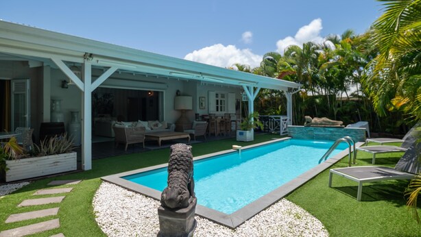 Piscina com uma escultura de pedra em primeiro plano, área de varanda coberta com móveis de exterior e vista para palmeiras ao fundo.