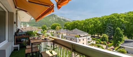 A balcony with a table and chairs, a view of a mountain, and a garden with various plants.