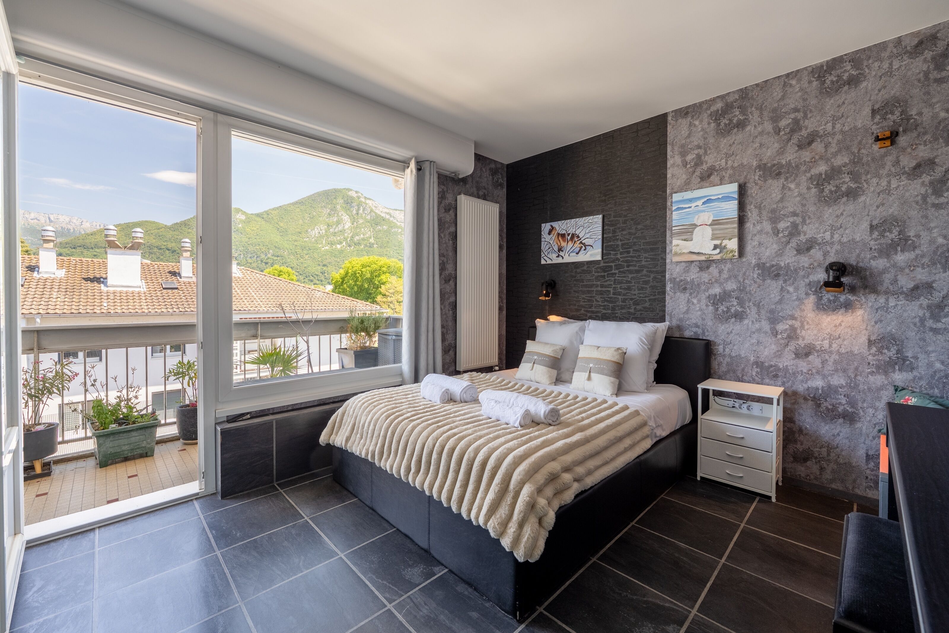 A modern bedroom with a large window, a bed with a fluffy white blanket, a nightstand, and two wall-mounted lamps.