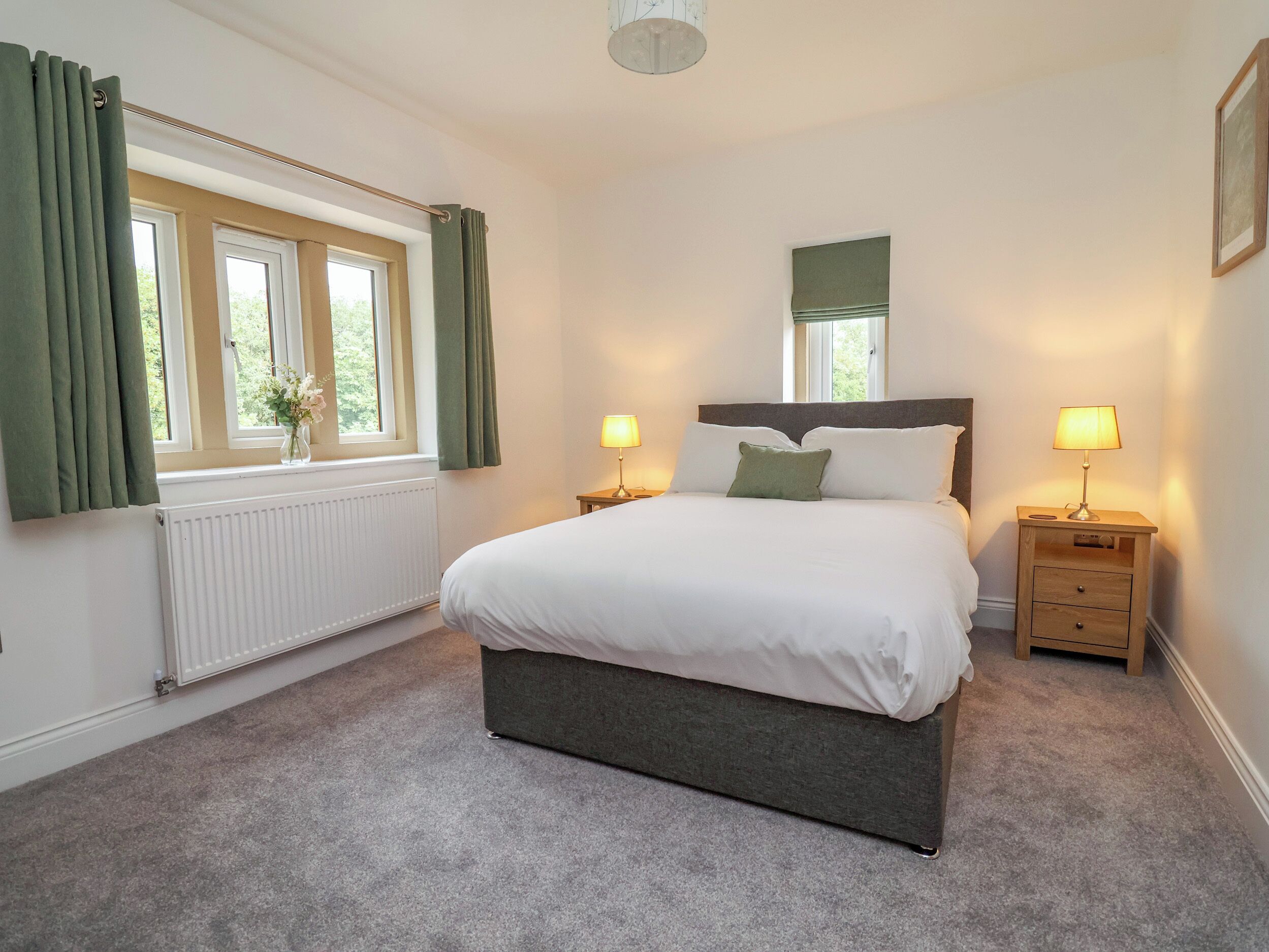 A bedroom with a bed, two bedside tables with lamps, a window with curtains, and a radiator.