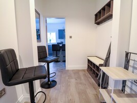 A narrow hallway with a seating area featuring two black chairs and a small table, leading to a kitchen area with blue cabinets and a window.